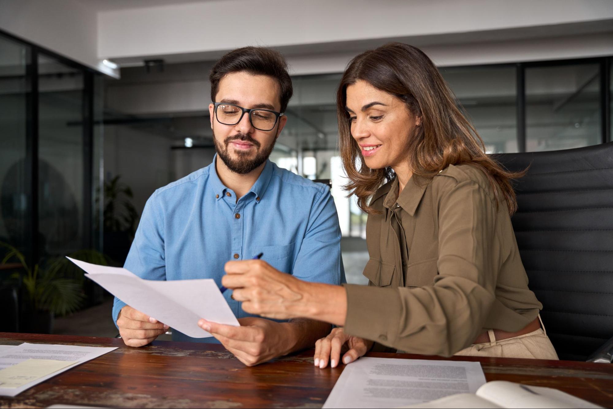 A man in a blue shirt and glasses and a woman in a brown blouse collaborate over negotiated trash bill at a wooden desk in a modern office, conveying teamwork.