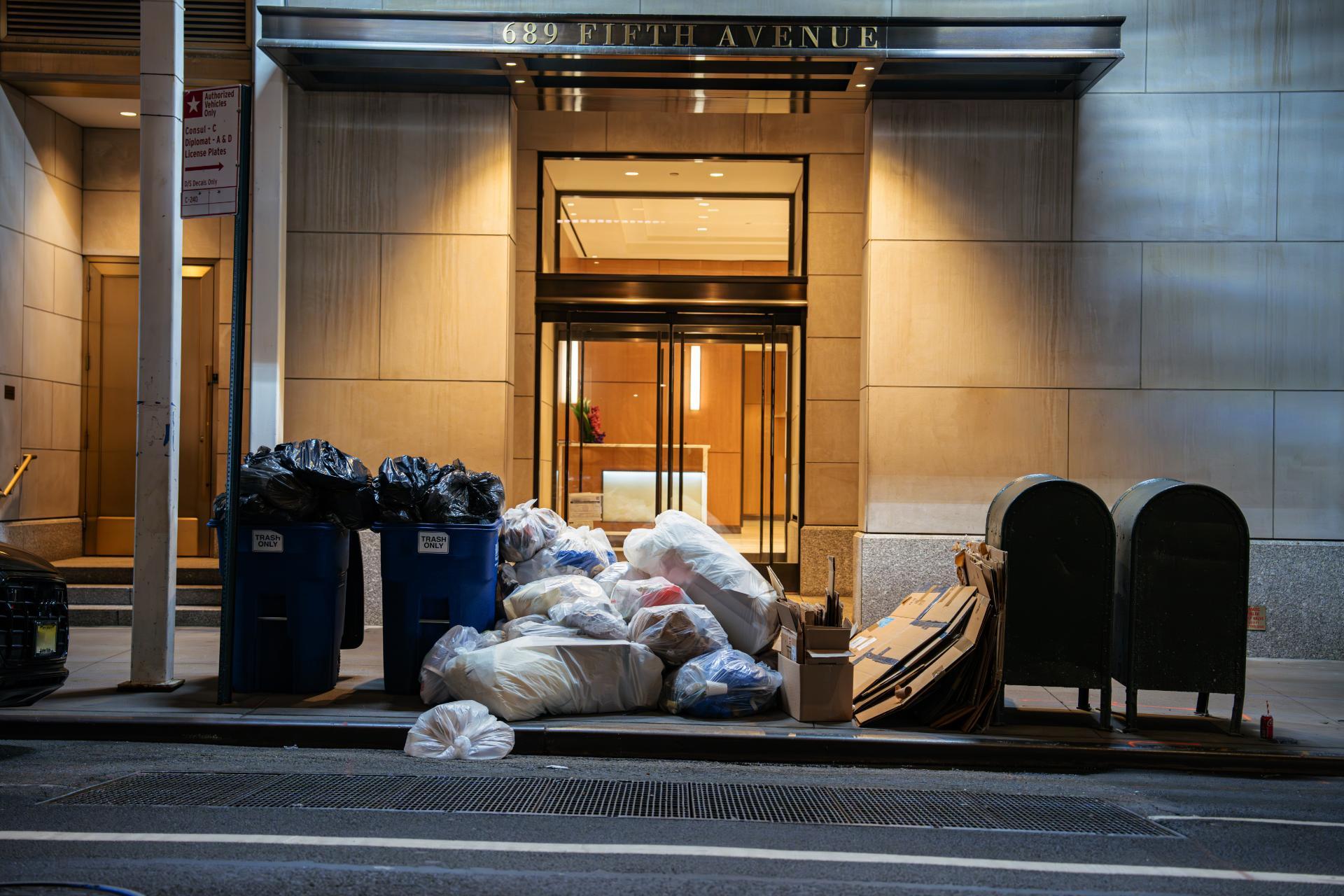 A pile of trash bags and cardboard boxes sits on the sidewalk outside a building entrance illuminated in soft light, creating an urban nighttime scene.