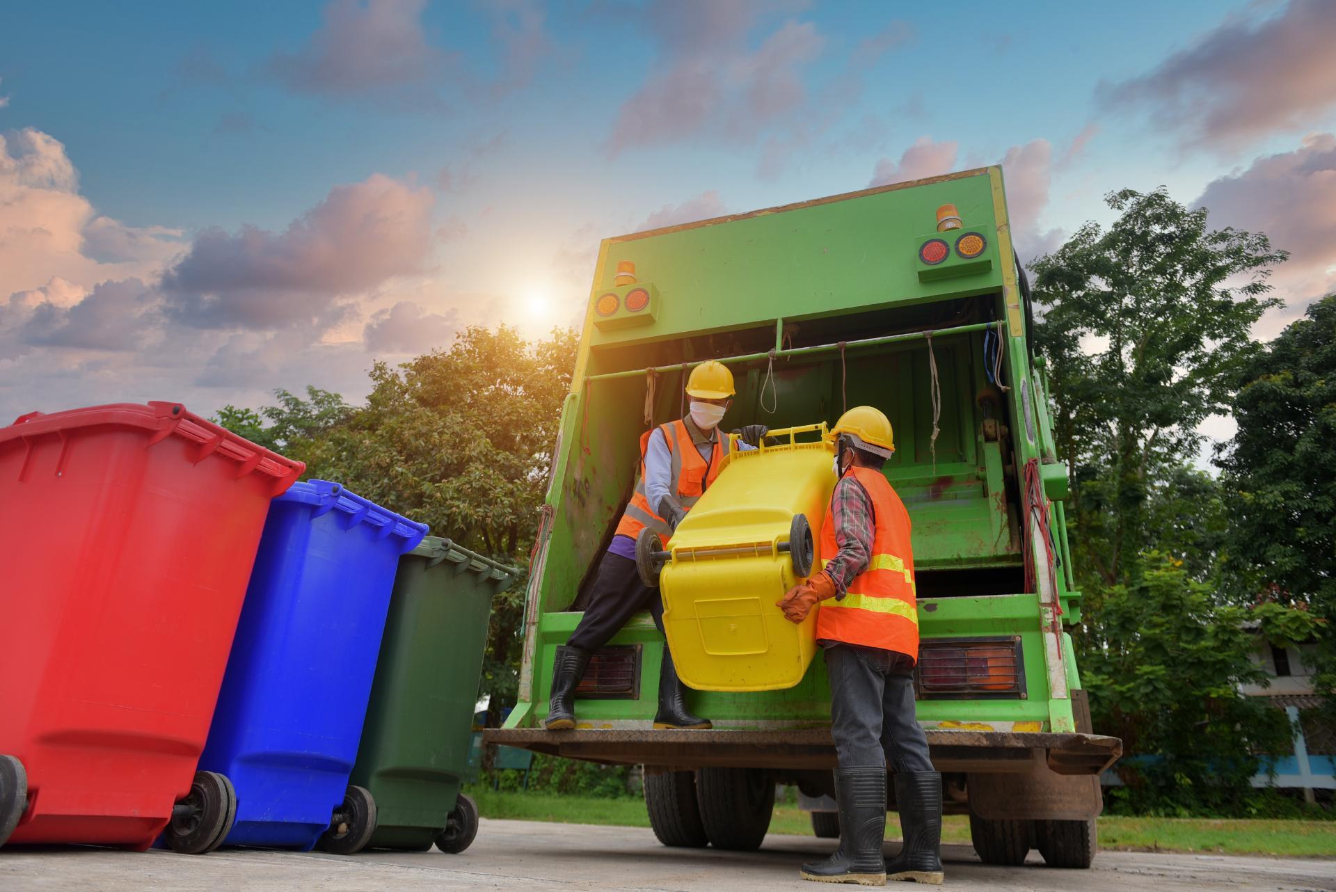 Sanitation workers in safety gear load a yellow bin into a green garbage truck at sunset, with red, blue, and green bins nearby. The mood is diligent and industrious.