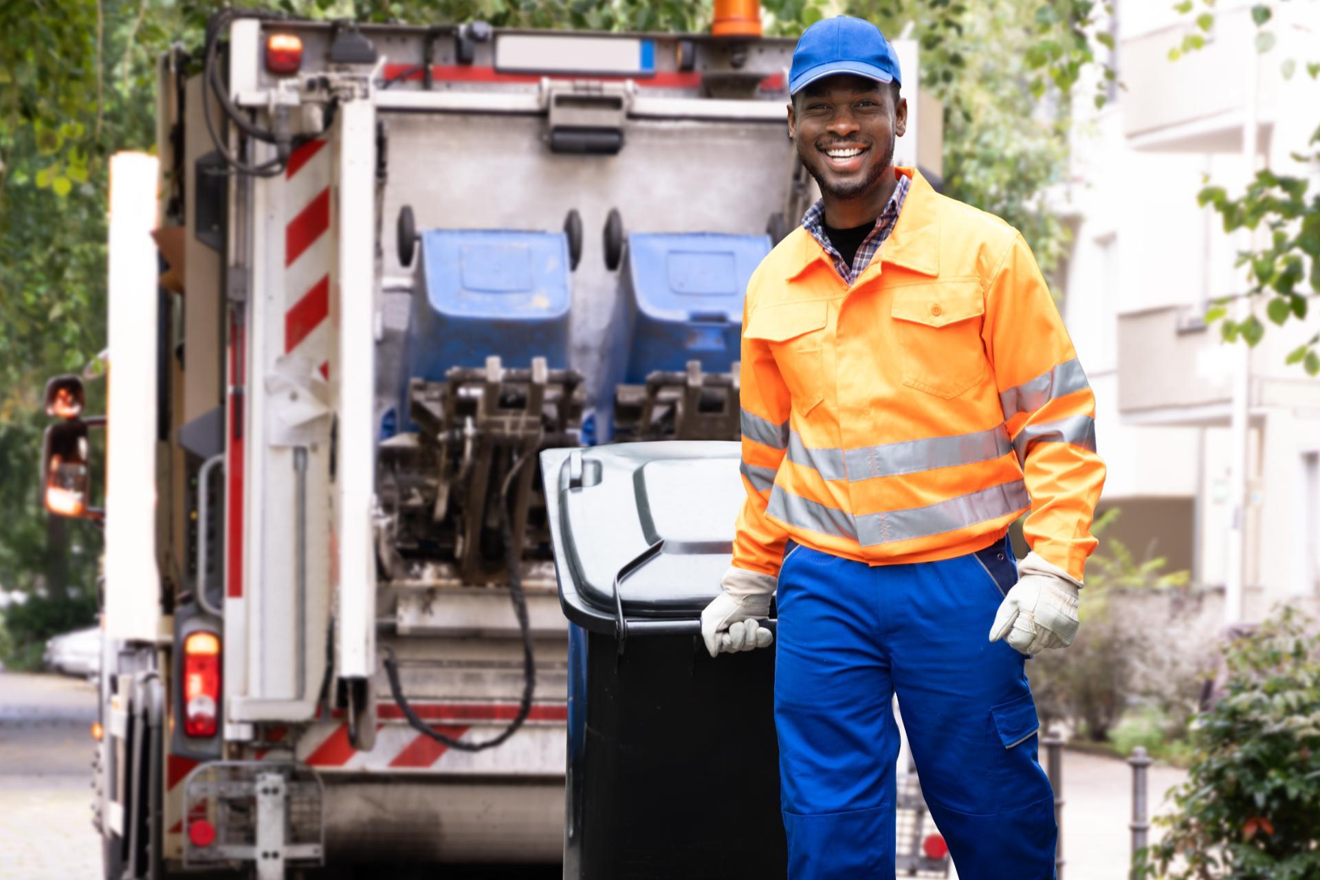 Smiling sanitation worker in bright orange and blue uniform stands beside a garbage truck holding a black trash bin. Trees and street in background.