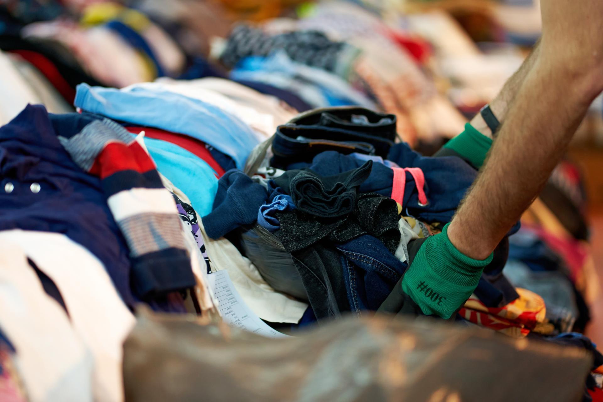 A person wearing green gloves sorts through a colorful pile of clothes to recycle.
