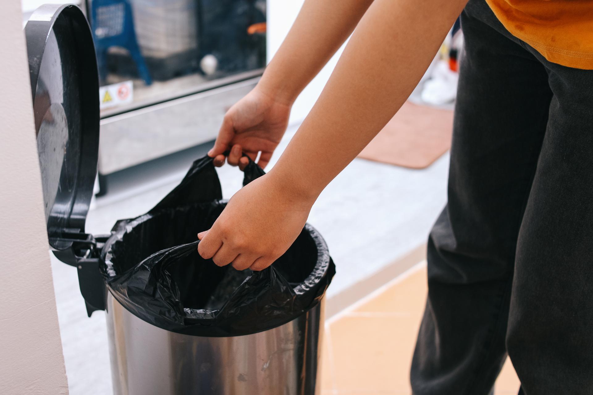A person places a black trash bag into a silver trash can in a clean, indoor setting. The image conveys tidiness and organization.