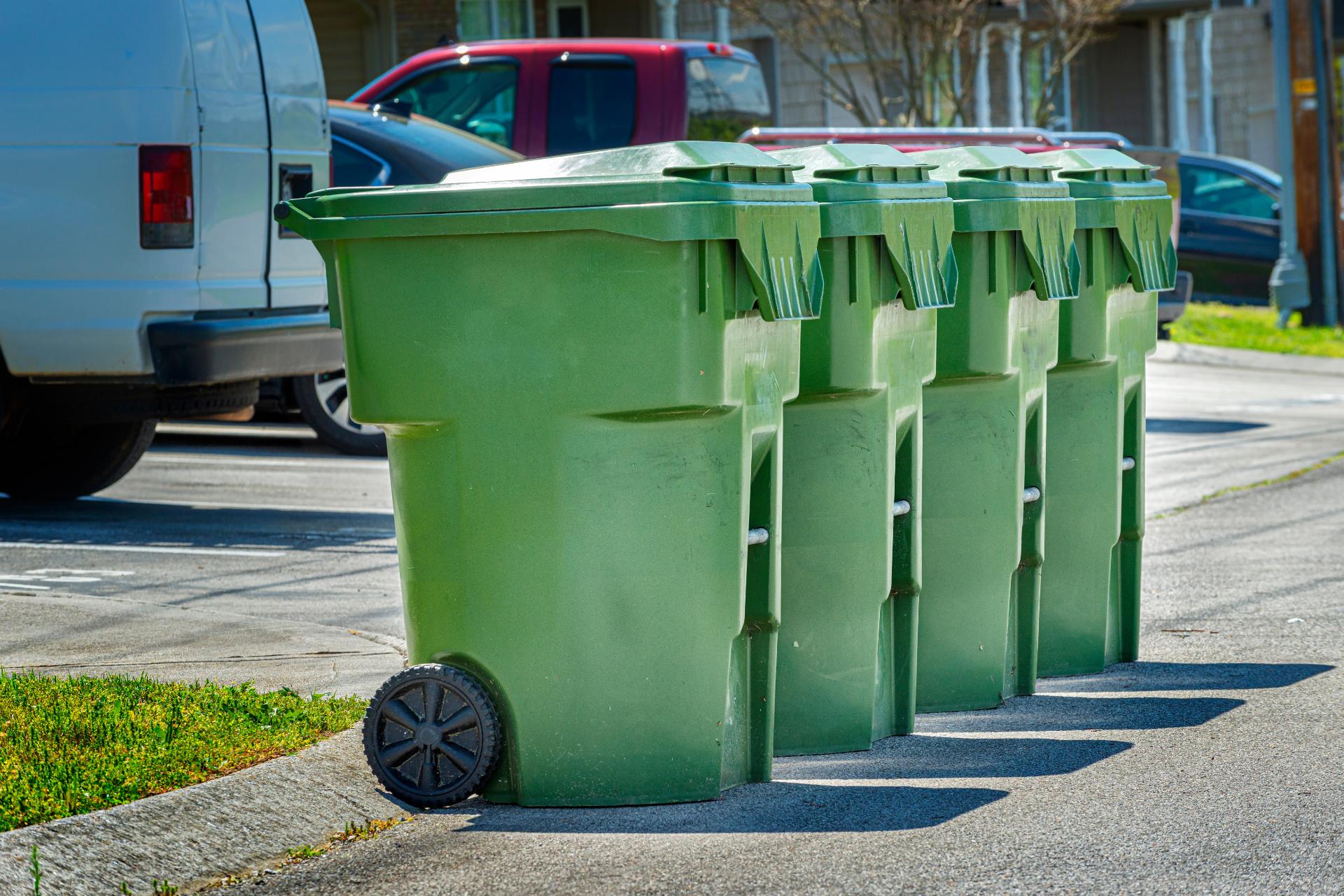 Four green recycling bins are lined up on a sunny street, next to parked cars. The atmosphere is calm, with bright lighting and clear skies.