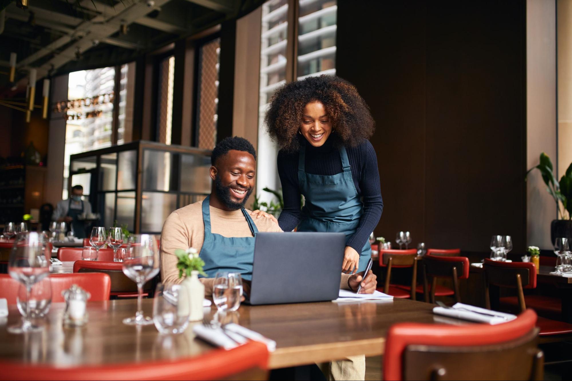 A man and a woman in a restaurant wearing aprons checking their waste metrics on a laptop